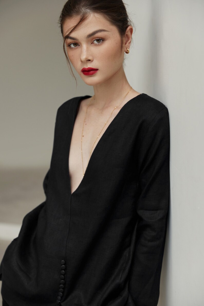 May include: A woman wearing a black, long-sleeved, v-neck top with a gold chain necklace. The top has a button closure and the woman is standing against a white wall.