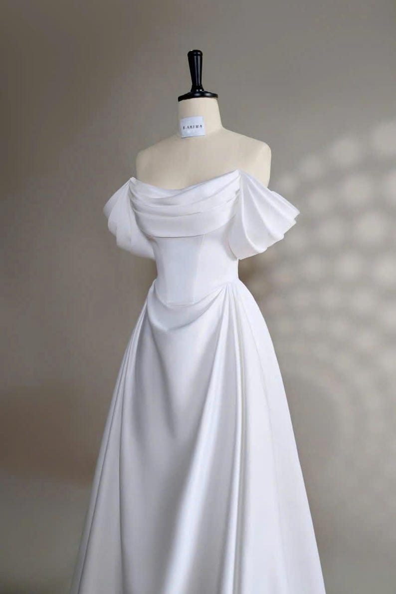 May include: A white off-the-shoulder wedding dress with a fitted bodice and a flowing skirt. The dress has a simple design with a slight ruffle detail on the shoulders. The dress is displayed on a mannequin against a light gray background.
