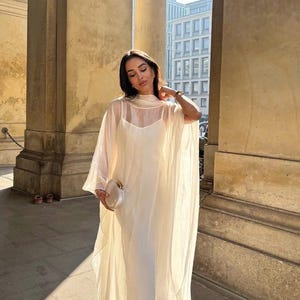 May include: A white sheer maxi dress with a high neck and a long, flowing skirt. The dress is layered over a white slip dress. The dress is being worn by a person standing in front of a building with columns.