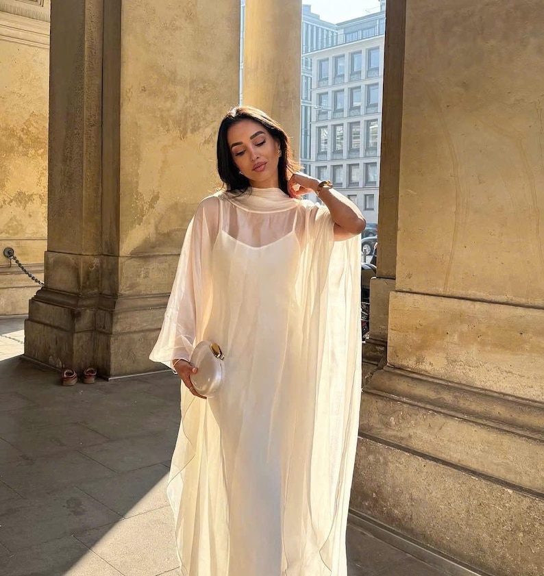 May include: A white sheer maxi dress with a high neck and a long, flowing skirt. The dress is layered over a white slip dress. The dress is being worn by a person standing in front of a building with columns.