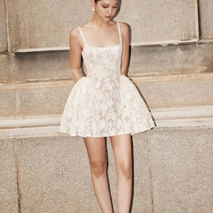 May include: A white lace mini dress with a square neckline and a fitted bodice. The dress has a flared skirt and is worn with white heels.
