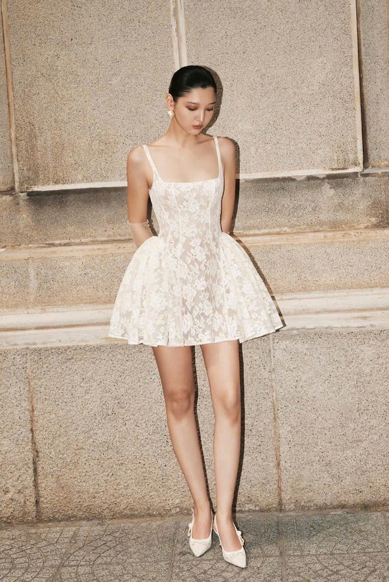 May include: A white lace mini dress with a square neckline and a fitted bodice. The dress has a flared skirt and is worn with white heels.