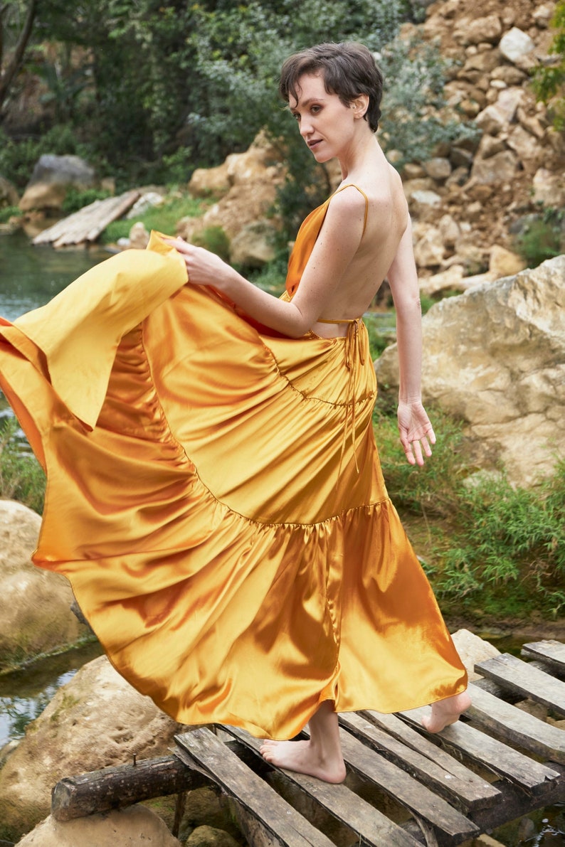 May include: A woman wearing a flowing, gold satin dress with a low back and a tiered skirt. She is standing on a wooden platform with a stream of water in the background.