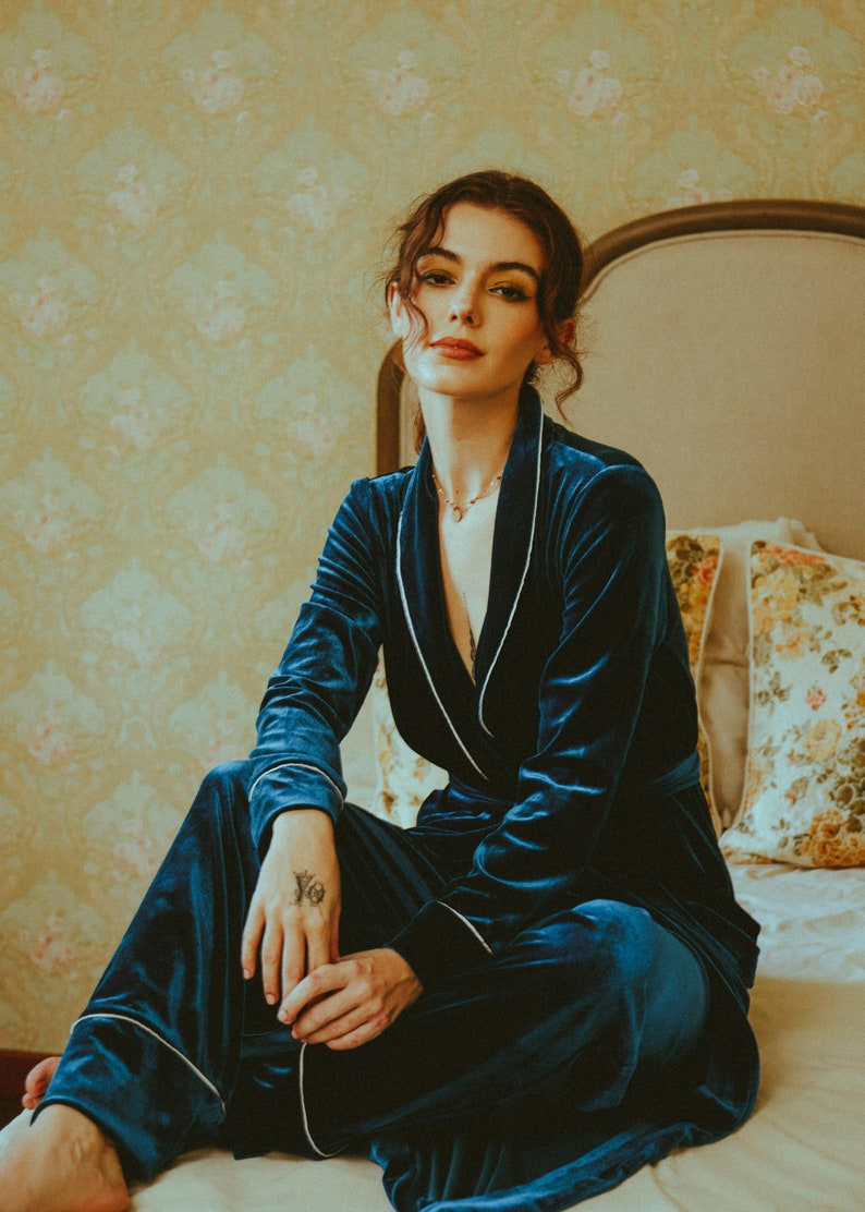 K&ouml;nnte beinhalten: Eine Frau tr&auml;gt eine Robe aus tealblauem Samt mit wei&szlig;en Paspeln. Sie sitzt auf einem Bett mit einem floral gemusterten Kissen hinter ihr.