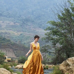 May include: A woman in a gold dress walks across a wooden bridge over a stream. The background is a lush green mountainside with fog.