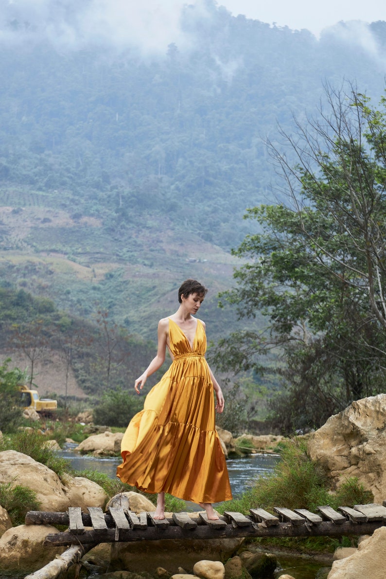 May include: A woman in a gold dress walks across a wooden bridge over a stream. The background is a lush green mountainside with fog.