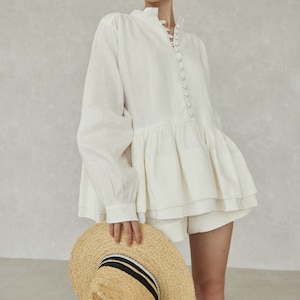May include: A white linen blouse with a ruffled hem and a button-up front. The blouse has long sleeves and a high neckline. The model is wearing the blouse with white shorts and a straw hat with a black and white striped band.