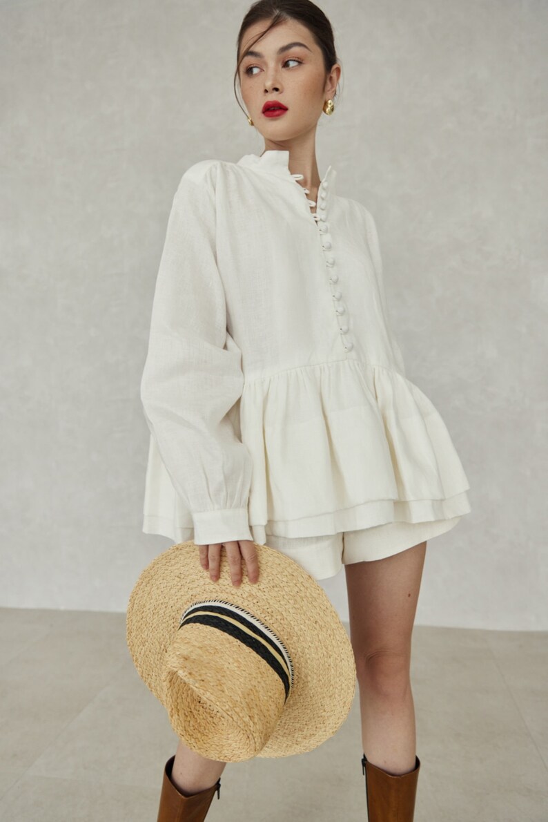 May include: A white linen blouse with a ruffled hem and a button-up front. The blouse has long sleeves and a high neckline. The model is wearing the blouse with white shorts and a straw hat with a black and white striped band.