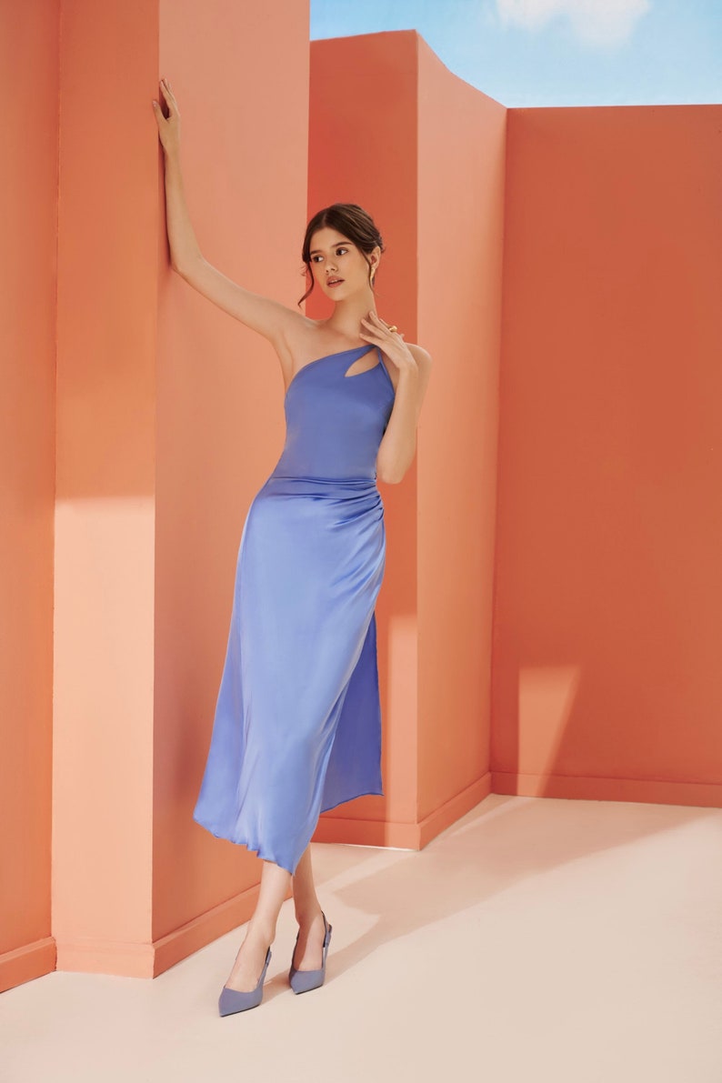 K&ouml;nnte beinhalten: Eine Frau in einem blauen Satin-Kleid mit einem Schultertr&auml;ger und einem Ausschnitt. Sie steht in einem Raum mit pfirsichfarbenen W&auml;nden.