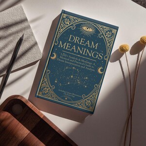 A styled overhead shot featuring the Dream Meanings book as part of a creative workspace. The hardcover book with its ornate blue-and-gold cover lies on a white table bathed in warm sunlight. To the left, a textured notebook and a pencil suggest a journaling moment , and to the right, a pair of yellow dried billy  Soft shadows stripe across the scene, enhancing the cozy yet mystical ambiance. This inviting composition shows the book as not just a reference guide but also a beautiful accessory
