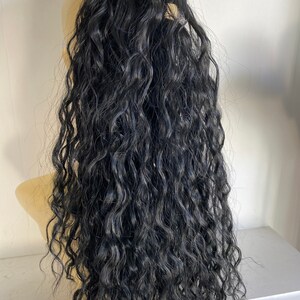 May include: A long, black, curly hair extension. The hair is thick and wavy, and it is tied up in a ponytail.