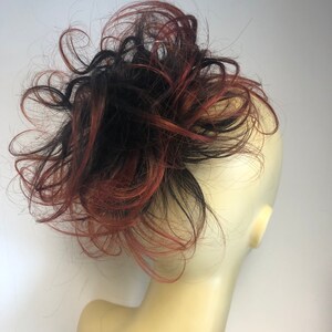 Könnte beinhalten: Ein unordentlicher Haarknoten-Haarteil mit einer Mischung aus schwarzen und rotbraunen Strähnen. Das Haarteil ist an einem Schaufensterkopf befestigt, um den Stil und die Farbvariationen zu zeigen. Der Knoten ist mit einem voluminösen, zerzausten Look gestylt.