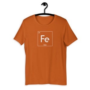 以下が含まれることがあります： オレンジ色の半袖Tシャツ。白い正方形のグラフィックには、鉄の化学記号「Fe」、原子番号「26」、そしてその下に「Iron」という文字が書かれています。