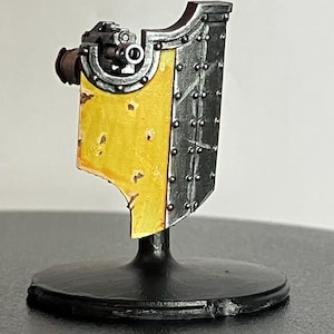 May include: A miniature metal shield with a yellow painted surface and a small gun mounted on the side. The shield is on a black display stand.