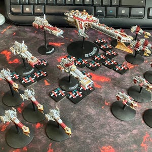 Może przedstawiać: Kolekcja pomalowanych miniaturowych modeli statków kosmicznych, zawierająca duży okręt flagowy i kilka mniejszych jednostek eskortujących. Statki są głównie białe, czerwone i złote, zamontowane na czarnych okrągłych podstawkach. Dołączono małe kwadratowe żetony.