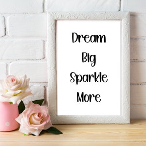 K&ouml;nnte beinhalten: Ein wei&szlig; gerahmter Druck mit den Worten &bdquo;Dream Big Sparkle More&ldquo; in schwarzer Schrift. Zwei rosa Rosen befinden sich in einer kleinen rosa Vase links vom Rahmen. Der Hintergrund ist eine wei&szlig;e Ziegelmauer und eine Holzoberfl&auml;che.