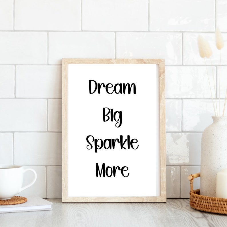 K&ouml;nnte beinhalten: Ein gerahmter Druck mit den Worten "Dream Big Sparkle More" in schwarzer Schrift. Der Rahmen ist aus hellem Holz, und der Druck wird auf einer wei&szlig;en Oberfl&auml;che mit einem wei&szlig;en Fliesen Hintergrund angezeigt. Eine wei&szlig;e Tasse und eine dekorative Vase sind ebenfalls sichtbar.