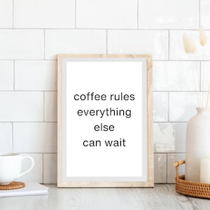Puede incluir: Un marco con la frase "coffee rules everything else can wait" en negro sobre fondo blanco. El marco es de color marrón claro. Se ve una taza de café blanca y una pequeña pila de libros.