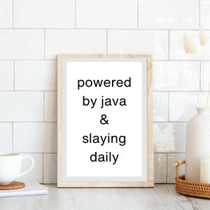 Puede incluir: Impresión enmarcada con las palabras en negro "powered by java & slaying daily". El marco es de color marrón claro. Una taza blanca y una pila de libros están a la izquierda. Un jarrón y una vela blancos están a la derecha.