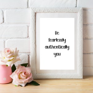 May include: A white framed print with the words "Be fearlessly authentically you" in black script. Two pink roses are in a pink vase to the left of the frame. The background is a white brick wall and a wooden surface.
