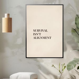 May include: A framed print with the text "SURVIVAL ISN'T ALIGNMENT" in a minimalist style. The print has a cream-coloured background and a black frame. The image is displayed on a wall with a neutral colour scheme.