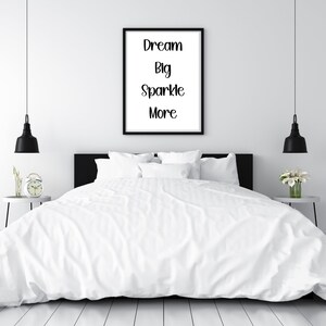 K&ouml;nnte beinhalten: Ein Schlafzimmer mit einem Bett mit wei&szlig;er Bettw&auml;sche und Kissen. Ein gerahmter Druck an der Wand zeigt die Worte "Dream Big Sparkle More". Schwarze Pendelleuchten h&auml;ngen auf beiden Seiten des Bettes. Ein Nachttisch mit einer Uhr und ein weiterer mit Blumen sind sichtbar.