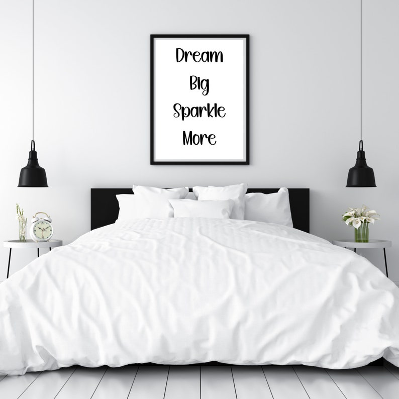K&ouml;nnte beinhalten: Ein Schlafzimmer mit einem Bett mit wei&szlig;er Bettw&auml;sche und Kissen. Ein gerahmter Druck an der Wand zeigt die Worte "Dream Big Sparkle More". Schwarze Pendelleuchten h&auml;ngen auf beiden Seiten des Bettes. Ein Nachttisch mit einer Uhr und ein weiterer mit Blumen sind sichtbar.