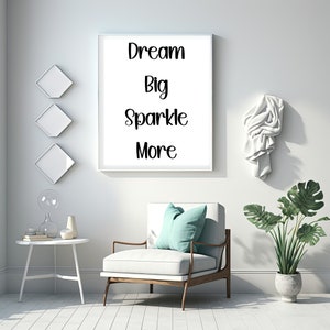 K&ouml;nnte beinhalten: Ein gerahmter Druck mit den Worten "Dream Big Sparkle More" h&auml;ngt an einer wei&szlig;en Wand. Darunter steht ein wei&szlig;er Sessel mit einem t&uuml;rkisfarbenen Kissen neben einem kleinen wei&szlig;en Tisch. Eine Topfpflanze und dekorative Wandkunst vervollst&auml;ndigen die Szene.
