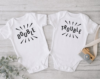 May include: Two white baby onesies with black text. One onesie says "DOUBLE" with radiating lines. The other onesie says "TROUBLE" with radiating lines.