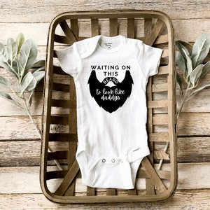 May include: White baby onesie with black text that reads "Waiting on this beard to look like daddy's". The onesie is in a wooden basket.