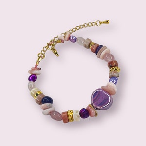 Könnte beinhalten: Ein goldfarbenes Kettenarmband mit einem herzförmigen Anhänger und verschiedenen rosa, violetten und weißen Perlen.