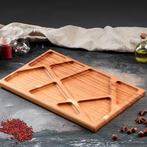 Puede incluir: Una bandeja de servir rectangular de madera con múltiples compartimentos triangulares. La bandeja está hecha de madera marrón claro y tiene un acabado natural.
