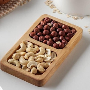 Puede incluir: Una bandeja de servir de madera con dos compartimentos, uno lleno de anacardos y el otro de avellanas.