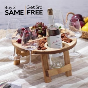 K&ouml;nnte beinhalten: Ein h&ouml;lzerner Picknicktisch mit vier Weingl&auml;sern aus Glas und einer Flasche Wein. Der Tisch hat einen integrierten Flaschenhalter und vier Schlitze f&uuml;r Gl&auml;ser. Der Tisch steht auf einer wei&szlig;-braunen gestreiften Decke. Der Text "Buy 2 SAME FREE" und "Get 3rd FREE" ist auf dem Bild zu sehen.
