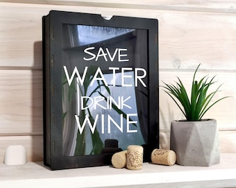 May include: A rectangular black wooden shadow box with a glass front featuring the white text SAVE WATER DRINK WINE. The box is accompanied by two natural corks, one with a grape vine illustration, and a small green plant in a gray geometric pot.