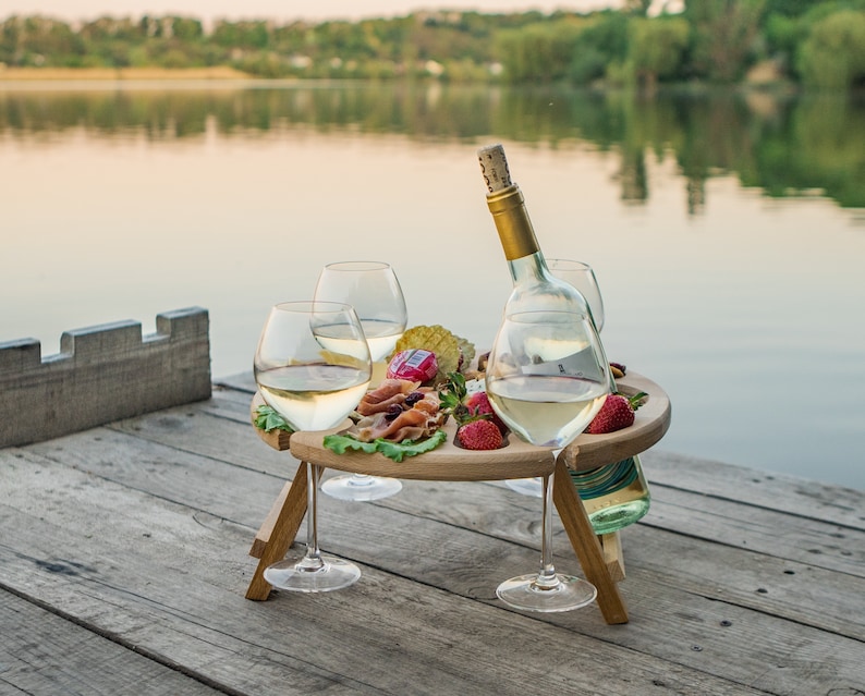 K&ouml;nnte beinhalten: Ein h&ouml;lzerner Picknicktisch mit einem integrierten Flaschenhalter und vier Weingl&auml;sern aus Glas. Der Tisch ist mit einem K&auml;se- und Wurstteller, Erdbeeren und einer Flasche Wei&szlig;wein gedeckt. Der Tisch steht auf einem Holzsteg mit Blick auf einen See.
