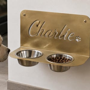 Peut inclure: Une mangeoire murale en métal doré avec deux bols circulaires en acier inoxydable. Le panneau arrière est découpé au laser avec le nom Charlie en écriture cursive et un motif de patte, fixé au mur avec deux vis métalliques visibles.