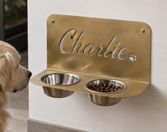 May include: A wall-mounted gold-toned metal pet feeder featuring two circular stainless steel bowls. The back panel is laser-cut with the name Charlie in a cursive script and a small paw print design, secured to the wall with two visible screws.