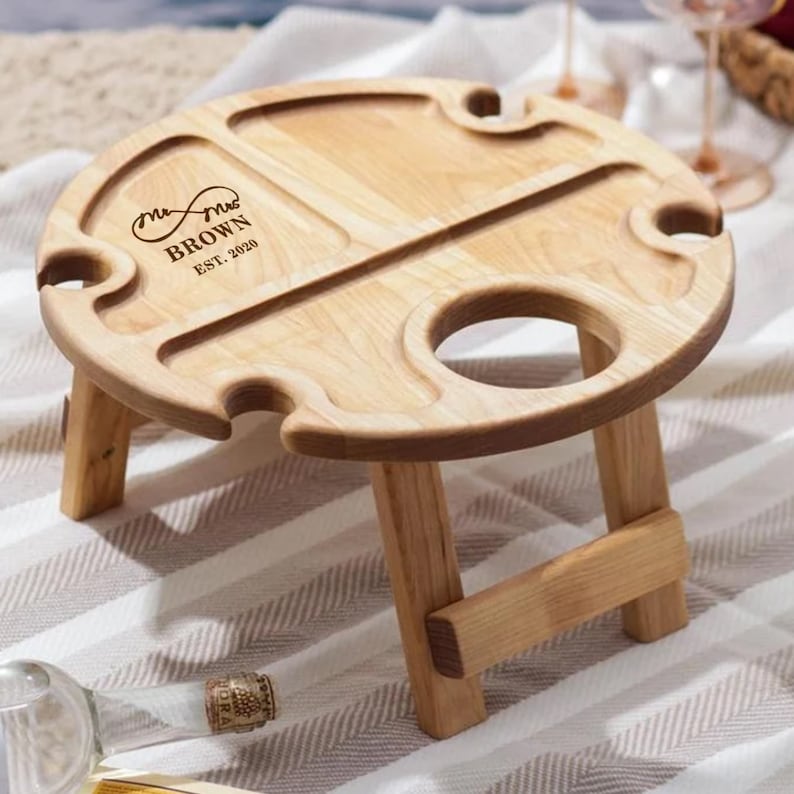 May include: A wooden picnic table with a round top and two folding legs. The table has a built-in bottle opener and two cup holders. The table is engraved with the text "Mr. & Mrs. Brown Est. 2020".