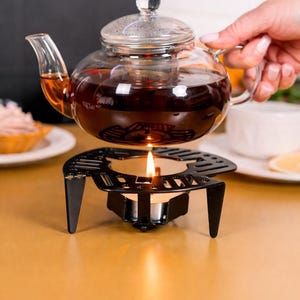 Könnte beinhalten: Eine Teekanne aus klarem Glas, gefüllt mit dunkel bernsteinfarbenem Tee, mit herausnehmbarem Siebeinsatz und Glasdeckel. Die Kanne steht auf einem schwarzen Metall-Stövchen, in dessen Mitte ein brennendes Teelicht die Temperatur des Tees hält.