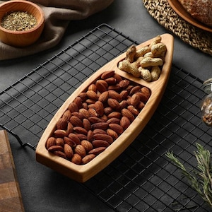 May include: A boat-shaped wooden serving tray with two distinct compartments. The larger section is filled with brown almonds, while the smaller, pointed section contains light brown peanuts in their shells. The tray has a smooth, light wood finish.