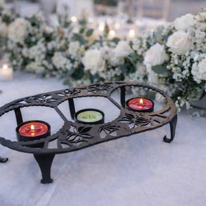 Könnte beinhalten: Ein schwarzer Kerzenhalter aus Metall mit ovalem Rahmen und filigranen floralen Ausschnitten, der auf vier geschwungenen Beinen steht. Der Halter enthält drei kleine Teelichter mit brennenden Flammen in rotem, grünem und rosa Wachs.