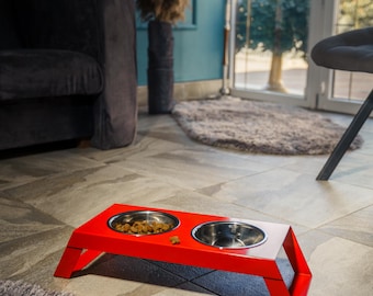 May include: A bright red elevated pet feeder stand featuring a geometric, angular frame with two circular cutouts. Two removable stainless steel bowls are inserted into the stand, with one bowl containing dry brown kibble pieces.