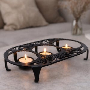 May include: A black metal candle holder with three lit tea light candles. The holder has an oval shape with decorative cut-out leaf patterns and short legs. The flames of the candles are bright against the dark metal.