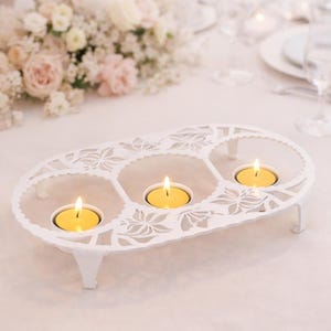 Può includere: Un portacandele in metallo bianco dalla forma ovale allungata con intricati intagli floreali. Il supporto presenta tre aperture circolari, ognuna contenente una piccola candela tea light rotonda con fiamma gialla, su quattro gambe corte.
