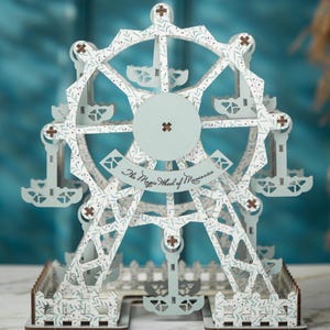 May include: A detailed, laser-cut wooden Ferris wheel model with a light blue and white color scheme. The wheel features intricate leaf patterns and the text "The Magic Wheel of Memories". The base is a detailed fence design.