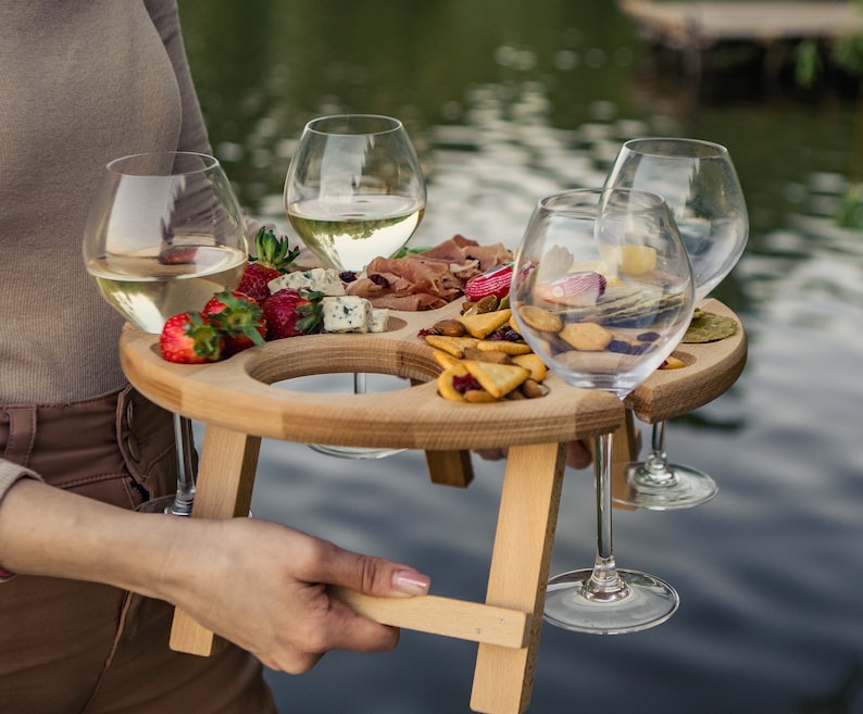 K&ouml;nnte beinhalten: Ein Holztablett mit vier Weingl&auml;sern aus Glas und einer Auswahl an Snacks, darunter Erdbeeren, K&auml;se und Cracker. Das Tablett hat einen eingebauten St&auml;nder und ist perfekt f&uuml;r die Unterhaltung im Freien.