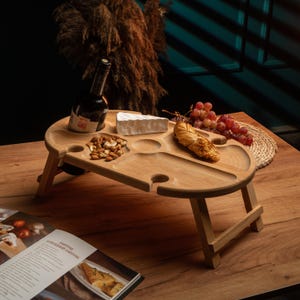 Puede incluir: Una mesa de picnic de madera con una botella de vino, queso, nueces y uvas. La mesa tiene huecos para copas de vino y comida, y está sostenida por patas plegables. La mesa es de madera clara.