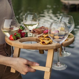 Könnte beinhalten: Ein rundes Serviertablett aus Holz mit vier integrierten kreisförmigen Aussparungen für Weingläser und einem vertieften Bereich in der Mitte für Speisen. Das Tablett hat vier kurze, klappbare Beine und zwei gefüllte Weingläser.