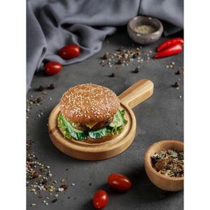 Op de afbeelding: Een burger op een sesambroodje met sla en komkommerschijfjes, geserveerd op een ronde houten serveerplank met handvat. De burger is voorzien van een tandenstoker en staat op een donkergrijs oppervlak met verspreide kruiden, tomaten en een kommetje.